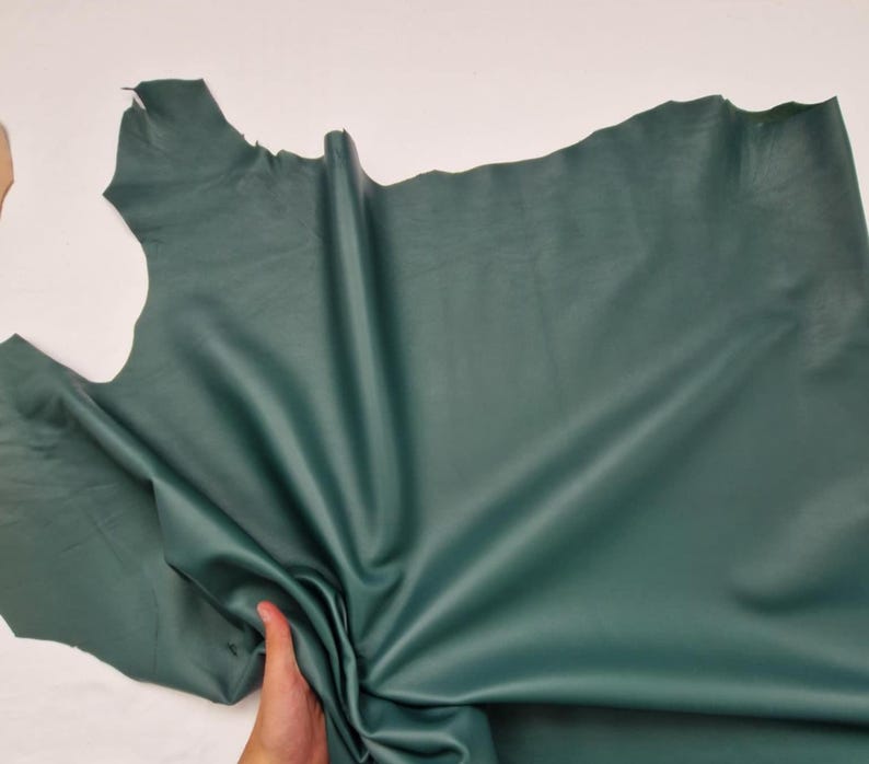 May include: A large piece of emerald green leather, with a smooth, matte finish. The leather is irregularly shaped, with natural edges. The material is held by a hand, showing the supple texture and potential for crafting. The color is consistent throughout the hide.