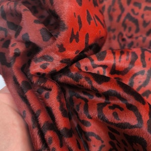 Op de afbeelding: Close-up van een rood-zwarte dierenprintstof. De stof heeft een zacht, getextureerd oppervlak met een patroon van zwarte vlekken en onregelmatige vormen tegen een rode achtergrond. Het materiaal is gevouwen, wat de textuur en diepte van de stof laat zien.