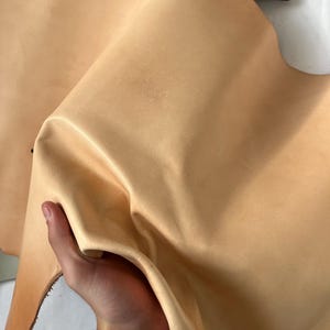 May include: A piece of light brown leather, folded and held in a hand. The leather has a smooth, slightly textured surface.