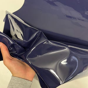 May include: A close-up of a dark blue, shiny, vinyl fabric. The fabric is folded and has a smooth, glossy finish.