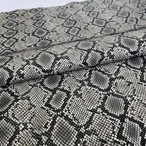 Könnte beinhalten: Nahaufnahme eines Stoffstücks mit Schlangenmuster. Das Muster ist ein sich wiederholendes Design aus grauen, schwarzen und weißen Schuppen. Der Stoff ist gefaltet und zeigt die Textur und die Details des Materials.