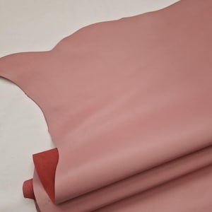May include: A stack of pink leather hides with a smooth texture. The top hide has a scalloped edge. A section of red leather is visible beneath the pink layers. The hides are likely for crafting or upholstery.