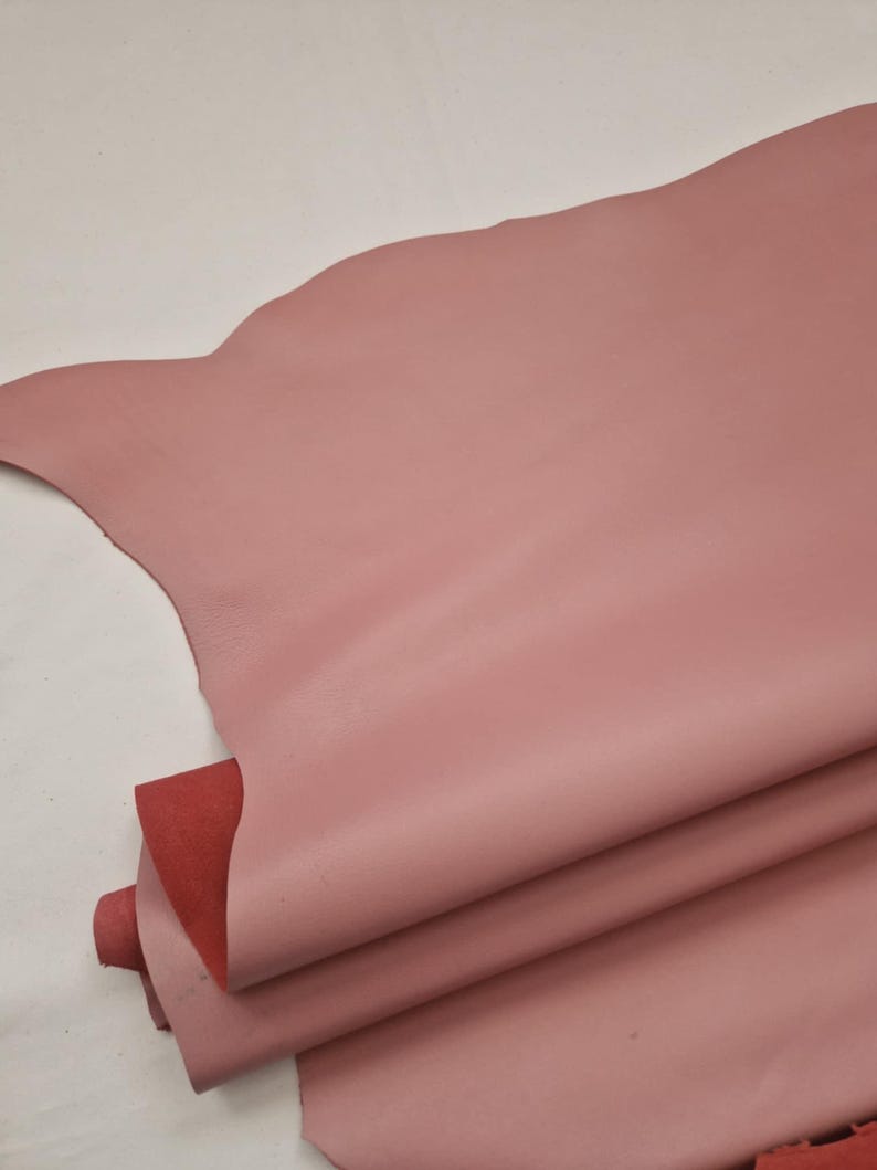 May include: A stack of pink leather hides with a smooth texture. The top hide has a scalloped edge. A section of red leather is visible beneath the pink layers. The hides are likely for crafting or upholstery.