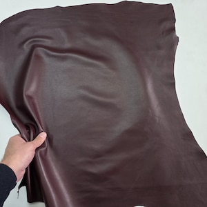 May include: A large piece of burgundy leather, held up to show its texture and flexibility. The leather has a smooth, slightly shiny surface and appears to be of high quality. The edges are uneven, indicating it's a cut piece, likely for crafting.