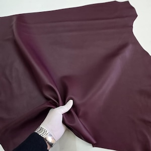 Puede incluir: Un gran trozo de cuero de color burdeos es sostenido por una mano enguantada. El cuero tiene una superficie lisa, ligeramente reflectante y parece flexible. Los bordes del cuero son irregulares, lo que sugiere que es un corte natural.