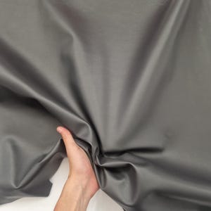 May include: A large sheet of gray leather with a smooth, shiny surface. The leather is being held up by a hand, showing its flexibility and texture. The material appears to be high-quality and suitable for various crafting projects.