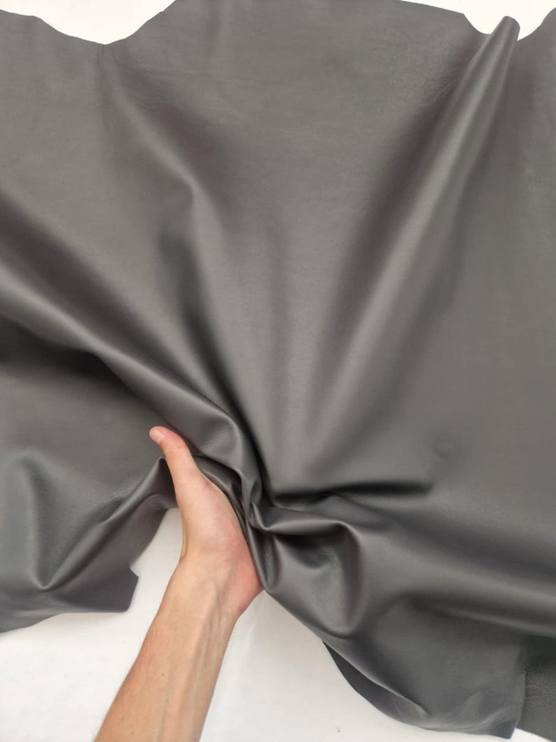 May include: A large sheet of gray leather with a smooth, shiny surface. The leather is being held up by a hand, showing its flexibility and texture. The material appears to be high-quality and suitable for various crafting projects.