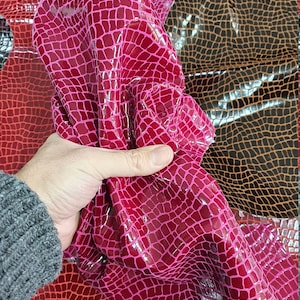 May include: A close-up of a pink faux leather fabric with a crocodile skin pattern. The fabric is being held in a hand and is folded over to show the texture.