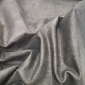 May include: Close-up of a piece of dark gray leather with a textured surface. The leather is folded and draped, showing its soft and supple texture.