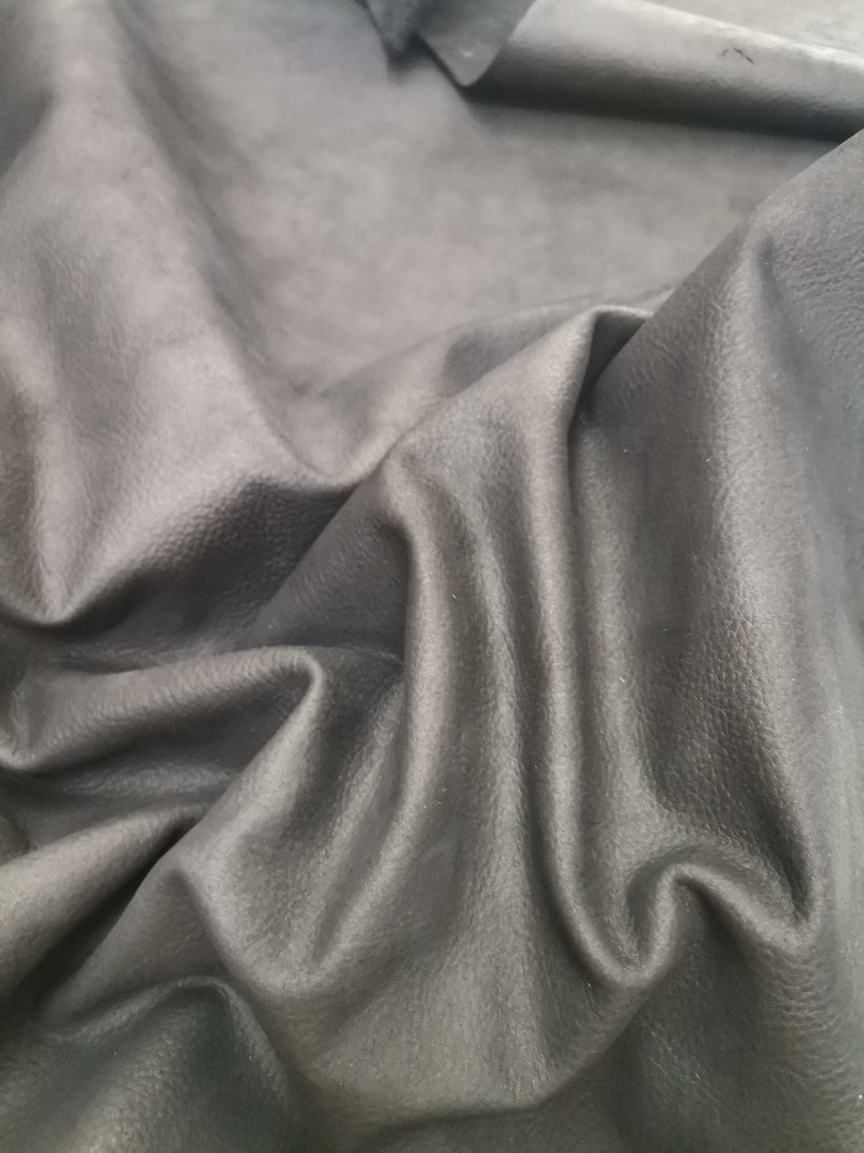 May include: Close-up of a piece of dark gray leather with a textured surface. The leather is folded and draped, showing its soft and supple texture.