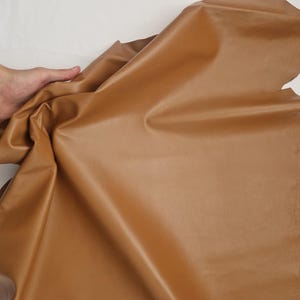 May include: A large piece of tan leather, with a smooth, slightly shiny surface. The leather is folded and draped, showing its flexibility. The edges are uneven, indicating a natural cut. The leather is a warm, neutral color.