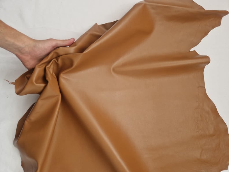 May include: A large piece of tan leather, with a smooth, slightly shiny surface. The leather is folded and draped, showing its flexibility. The edges are uneven, indicating a natural cut. The leather is a warm, neutral color.