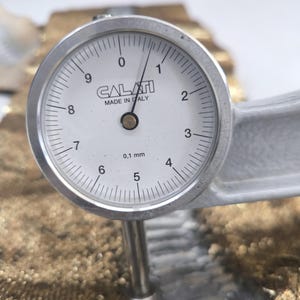 Pode incluir: Close-up de um medidor de mostrador prateado medindo uma superfície texturizada. O medidor, marcado "CALATI MADE IN ITALY", exibe uma medição de aproximadamente 0,1 mm. O fundo apresenta um material áspero marrom-dourado.