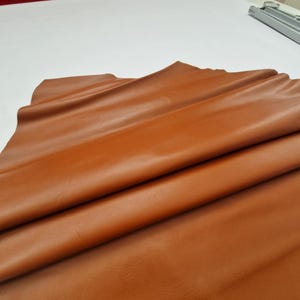 May include: A folded sheet of smooth, tan-colored leather. The leather has a glossy finish and appears to be supple. The folds create shadows and highlights, emphasizing the texture and color variations. The leather is likely used for crafting.