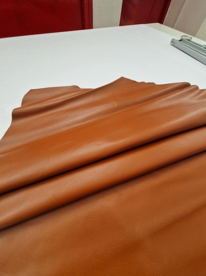 May include: A folded sheet of smooth, tan-colored leather. The leather has a glossy finish and appears to be supple. The folds create shadows and highlights, emphasizing the texture and color variations. The leather is likely used for crafting.