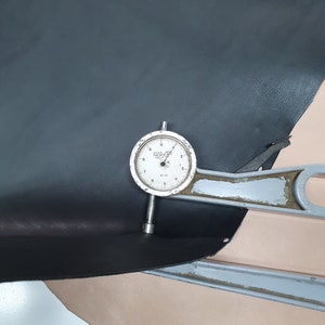 Puede incluir: Un primer plano de un medidor de cuero con un dial que marca 0,7 mm. El medidor est&aacute; apoyado en un trozo de cuero negro y un trozo de cuero rosa claro.