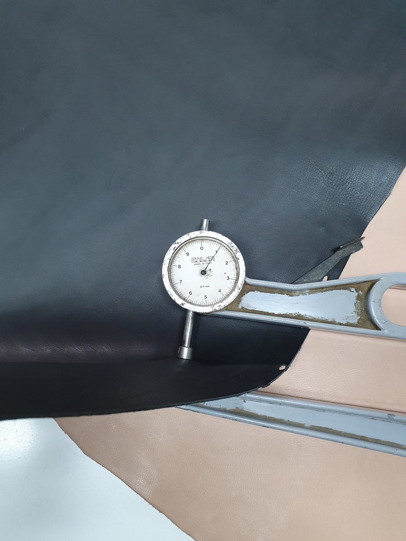 Puede incluir: Un primer plano de un medidor de cuero con un dial que marca 0,7 mm. El medidor est&aacute; apoyado en un trozo de cuero negro y un trozo de cuero rosa claro.