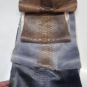 Peut inclure: Quatre morceaux de cuir de serpent dans différentes couleurs : marron, beige, gris et noir. Le cuir est disposé en pile, avec le morceau marron sur le dessus et le morceau noir en bas.