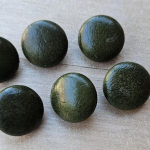 May include: Six dark green, round cabinet knobs are arranged on a light wood surface. The knobs have a slightly textured appearance and a glossy finish. These decorative knobs could be used for furniture or other home decor projects.