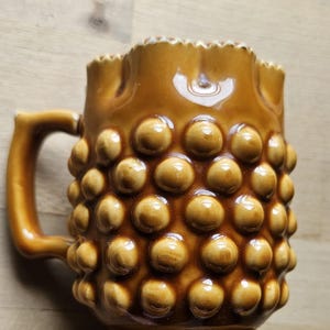 Op de afbeelding: Een bruine keramische mok met een textuur, bubbelachtig patroon. De mok heeft een handvat en een geschulpte rand. De mok heeft een warme, aardse tint en is waarschijnlijk vintage of retro.