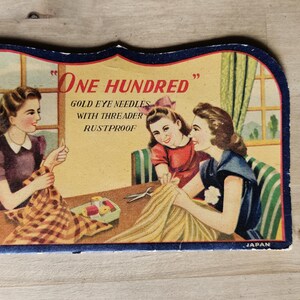 May include: Vintage needle tin with a retro illustration of three women sewing. The tin features the text "One Hundred Gold Eye Needles with Threader Rustproof". The image has a blue border and is set against a light background.