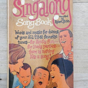 以下が含まれることがあります： レトロな表紙のヴィンテージ「The Lancer Singalong Song Book」。ピンク色のタイトルが際立ち、歌っている人々のイラストが描かれています。この本は「あなたのALL-TIMEお気に入りの曲の数々の言葉と音楽」を約束しています。