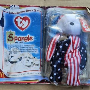 May include: A TY Beanie Baby plush toy named "Spangle the Bear" in a plastic package. The bear is white with a red, white, and blue American flag design. The packaging includes text about the bear and the McDonald's logo.