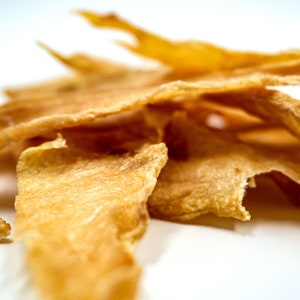May include: A pile of thin, crispy, light brown dog treats. The treats are made of dehydrated chicken and are shaped like long strips.