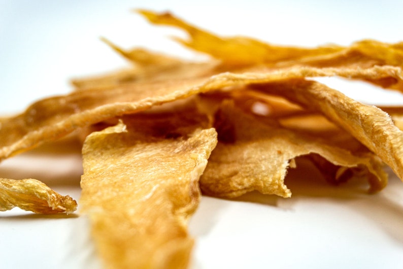 May include: A pile of thin, crispy, light brown dog treats. The treats are made of dehydrated chicken and are shaped like long strips.