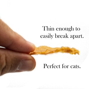 May include: A thin, light brown, dried meat treat for cats. The treat is being held by a hand and is thin enough to easily break apart.