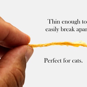 Puede incluir: Un premio de carne seca delgado y de color marr&oacute;n claro se sostiene entre dos dedos. El premio es lo suficientemente delgado como para romperse f&aacute;cilmente. El texto "Lo suficientemente delgado como para romperse f&aacute;cilmente. Perfecto para gatos." est&aacute; debajo de la imagen.