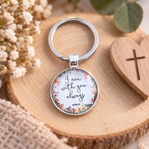 Könnte beinhalten: Ein runder Schlüsselanhänger aus silberfarbenem Metall mit einem Glaskabochon-Einsatz. Der Einsatz zeigt einen floralen Rand in Rosa- und Grüntönen, der den schwarzen Text "I am with you always" und die Referenz "Matthew 28:20" umrahmt.