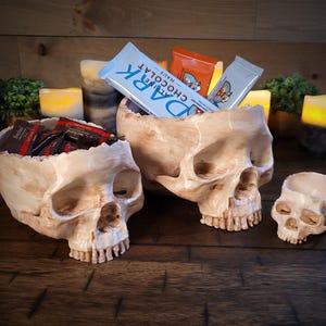 May include: Three skull-shaped containers, one small and two larger, are displayed on a wooden surface. The larger skulls hold candy and chocolate bars. The text "DARK CHOCOLAT" is visible on one of the chocolate bars.
