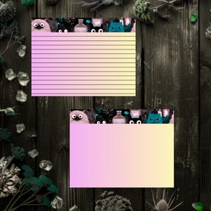 May include: Two rectangular note cards featuring a gradient background transitioning from pink to yellow. The top border of each card displays a dark illustration of various cats, potion bottles, and foliage. One card contains horizontal black lines.
