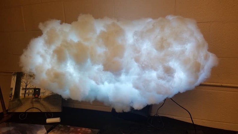 May include: A white cloud-shaped lamp with a white light shining through it. The lamp is made of cotton and is sitting on a black base.