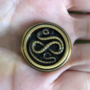 Puede incluir: Un botón redondo de metal en tono latón antiguo con un diseño de serpiente enroscada en relieve en el centro. La serpiente se muestra con detalles corporales segmentados y una cabeza circular, sobre un fondo oscuro y hundido con borde liso.