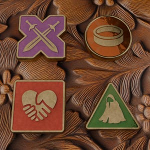 Puede incluir: Cuatro pines decorativos de metal con bordes dorados. Arriba a la izquierda, una forma de X morada con espadas cruzadas. Arriba a la derecha, un pin circular naranja con un icono de anillo. Abajo a la izquierda, un cuadrado rojo con un icono de apretón de manos. Abajo a la derecha, un triángulo verde con un icono de capa con capucha.