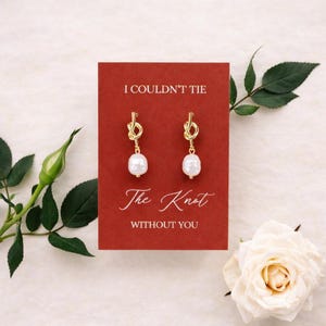 May include: Gold-tone knot earrings with a white pearl drop, displayed on a red card. The card reads "I Couldn't Tie The Knot Without You" in elegant script. A white rose and greenery frame the card.