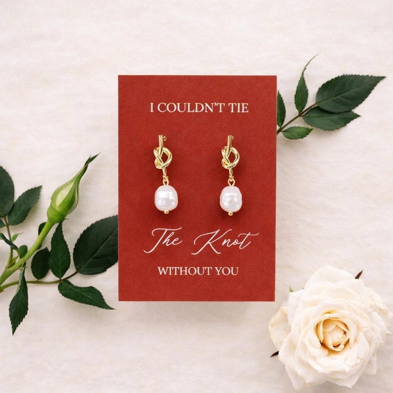 May include: Gold-tone knot earrings with a white pearl drop, displayed on a red card. The card reads "I Couldn't Tie The Knot Without You" in elegant script. A white rose and greenery frame the card.