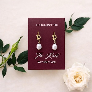 May include: Gold-colored knot earrings with white pearl drops are displayed on a burgundy card. The card reads "I COULDN'T TIE The Knot WITHOUT YOU" in white script. A white rose and green foliage frame the card.