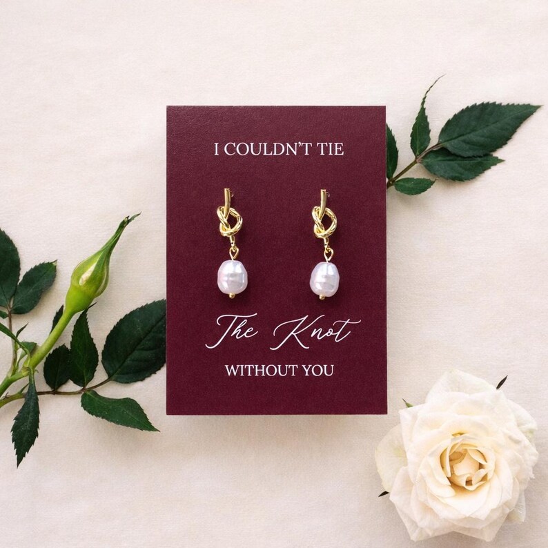 May include: Gold-colored knot earrings with white pearl drops are displayed on a burgundy card. The card reads "I COULDN'T TIE The Knot WITHOUT YOU" in white script. A white rose and green foliage frame the card.