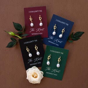 May include: Four pairs of gold-colored knot earrings with white pearl drops, displayed on cards of burgundy, navy, black, and green. Each card has the text "I COULDN'T TIE The Knot WITHOUT YOU". A white rose and greenery are also present.
