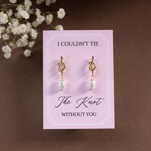 May include: Gold-tone knot earrings with a dangling white pearl, displayed on a pink card. The card reads "I Couldn't Tie The Knot Without You" in elegant script. White flowers are in the background.
