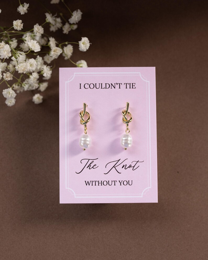 May include: Gold-tone knot earrings with a dangling white pearl, displayed on a pink card. The card reads "I Couldn't Tie The Knot Without You" in elegant script. White flowers are in the background.