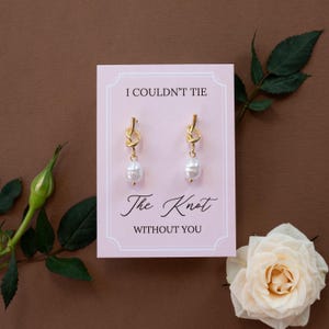 May include: Gold-colored knot earrings with a white pearl drop, displayed on a pink card with the text "I COULDN'T TIE The Knot WITHOUT YOU". The earrings are set against a brown background, with a white rose and green leaves.