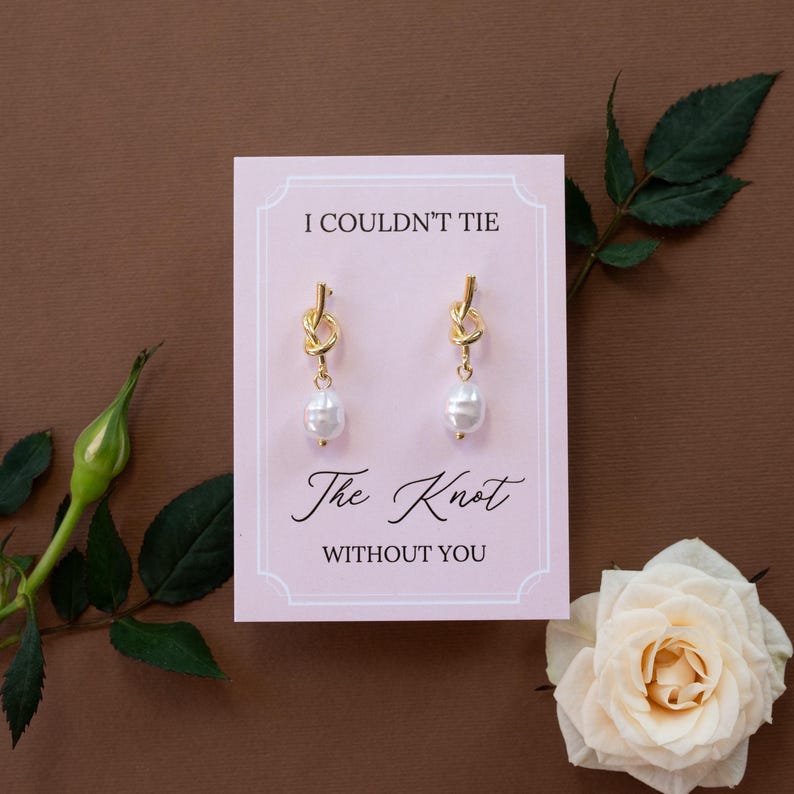 May include: Gold-colored knot earrings with a white pearl drop, displayed on a pink card with the text "I COULDN'T TIE The Knot WITHOUT YOU". The earrings are set against a brown background, with a white rose and green leaves.
