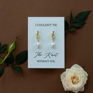 May include: Gold-colored knot earrings with a dangling white pearl, displayed on a card with the text "I COULDN'T TIE The Knot WITHOUT YOU". The card is on a brown background, with a white rose and green leaves.