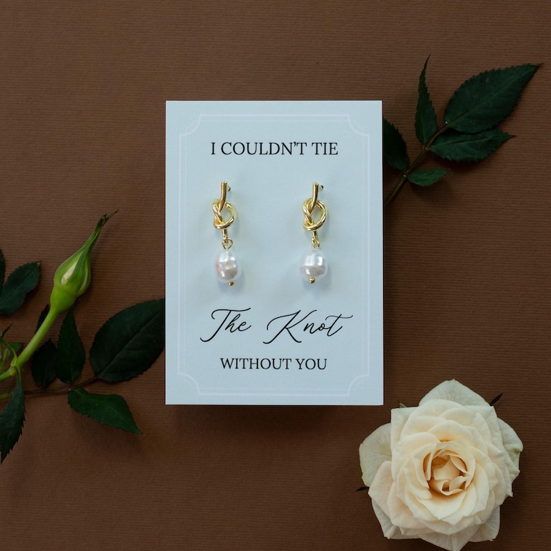 May include: Gold-colored knot earrings with a dangling white pearl, displayed on a card with the text "I COULDN'T TIE The Knot WITHOUT YOU". The card is on a brown background, with a white rose and green leaves.