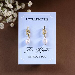 May include: Gold-colored knot earrings with a dangling pearl, presented on a light blue card. The card reads "I Couldn't Tie The Knot Without You" in elegant script. White flowers are in the background.