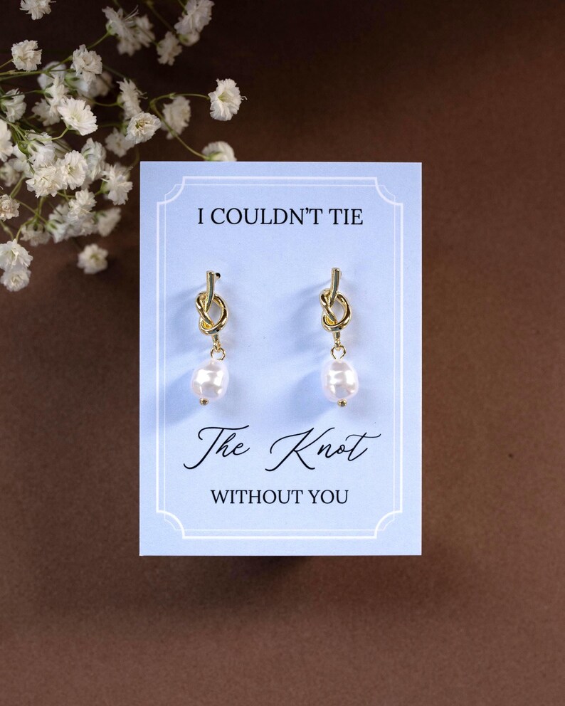 May include: Gold-colored knot earrings with a dangling pearl, presented on a light blue card. The card reads "I Couldn't Tie The Knot Without You" in elegant script. White flowers are in the background.